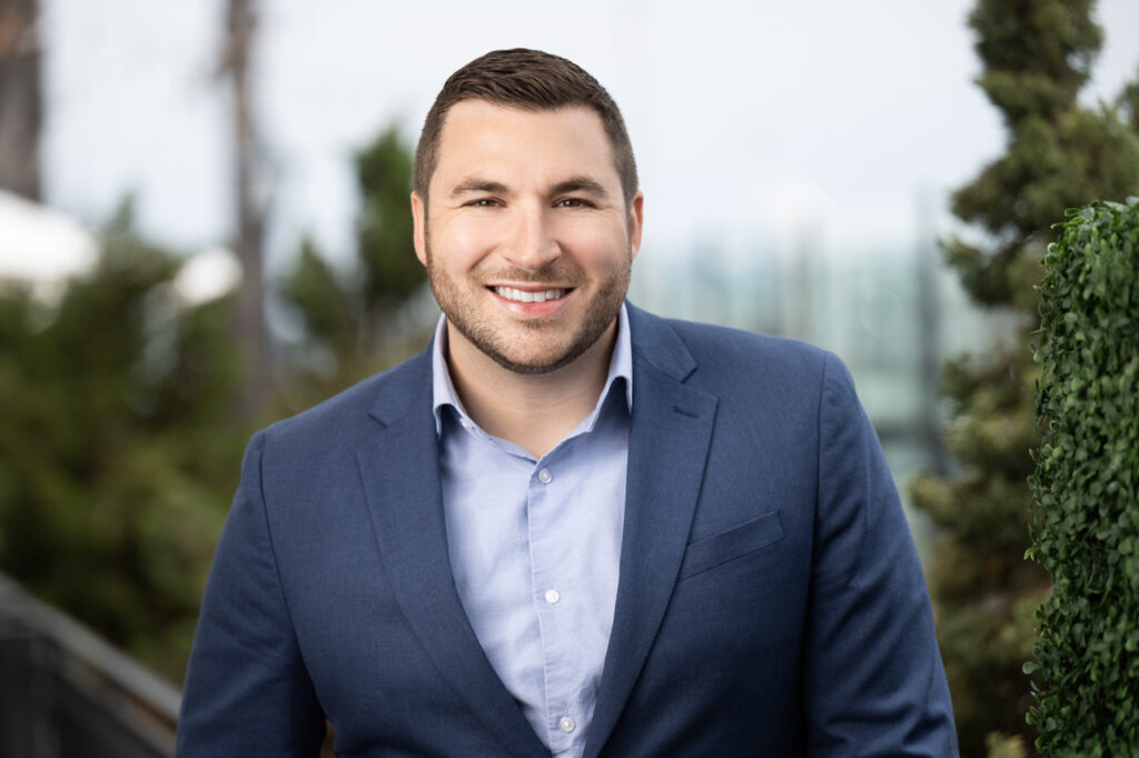 A person with short brown hair and a beard, wearing a blue blazer and light blue shirt, smiles while standing outdoors. The background is slightly blurred with greenery and structures visible, hinting at his success in project finance.