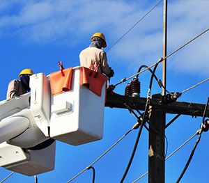 Two workers wearing helmets and harnesses are shown in elevated buckets repairing electrical power lines against a bright blue sky. Utilizing equipment finance, they are performing maintenance on a utility pole using the specialized tools from their bucket trucks.