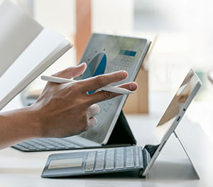 A person holding a stylus points at a project finance graph displayed on a tablet screen. Another tablet with a keyboard is positioned nearby on the white desk, alongside some documents and a book partially visible. Bright natural light illuminates the workspace.