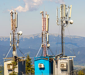 Three telecommunications towers with various antennas and equipment, financed through project finance initiatives, stand on a rooftop against a background of rolling mountains and a partly cloudy sky. The towers are equipped with different arrays of satellite dishes and antennas.